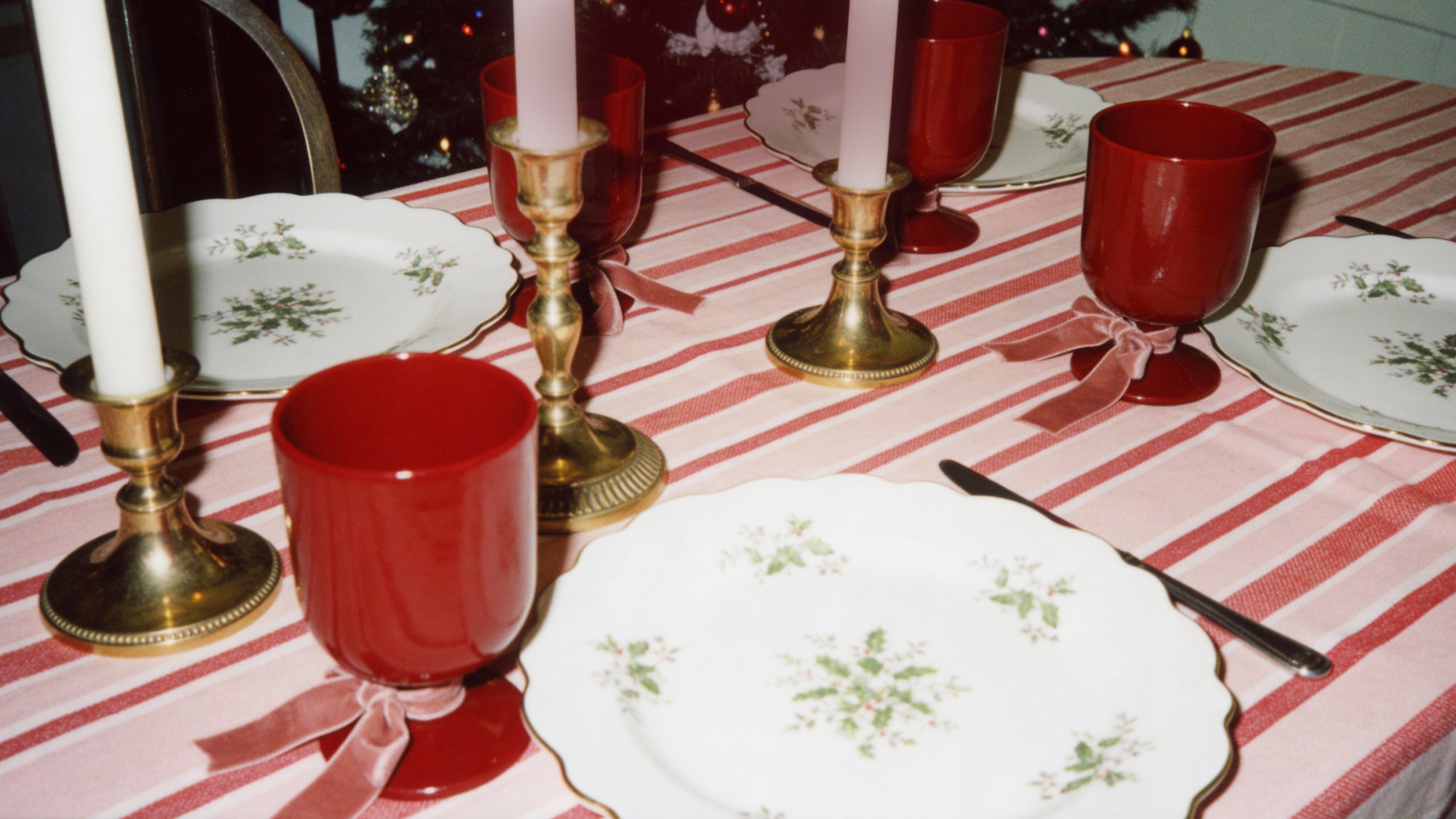 Dining table set with red burgundy Jarvise ceramic glasses, gold candle holders, and white plates with floral patterns on a pink and red striped tablecloth.